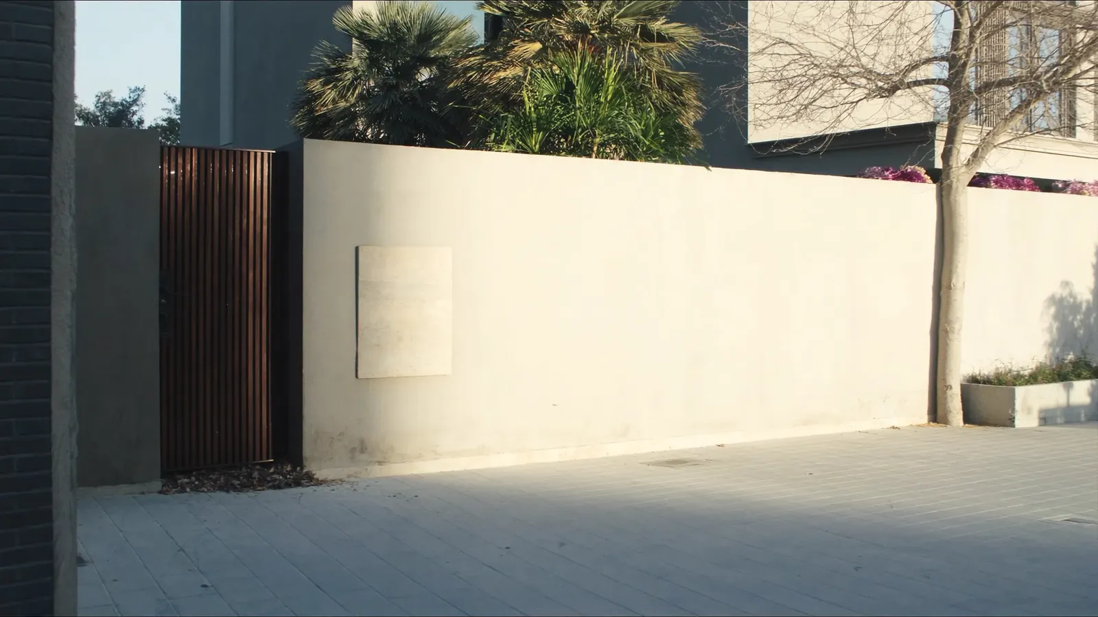 The finished street elevation of a contemporary Dubai villa in low afternoon sun: rendered boundary wall, a bronze slatted gate, a blank stone plaque set into the wall and planting behind