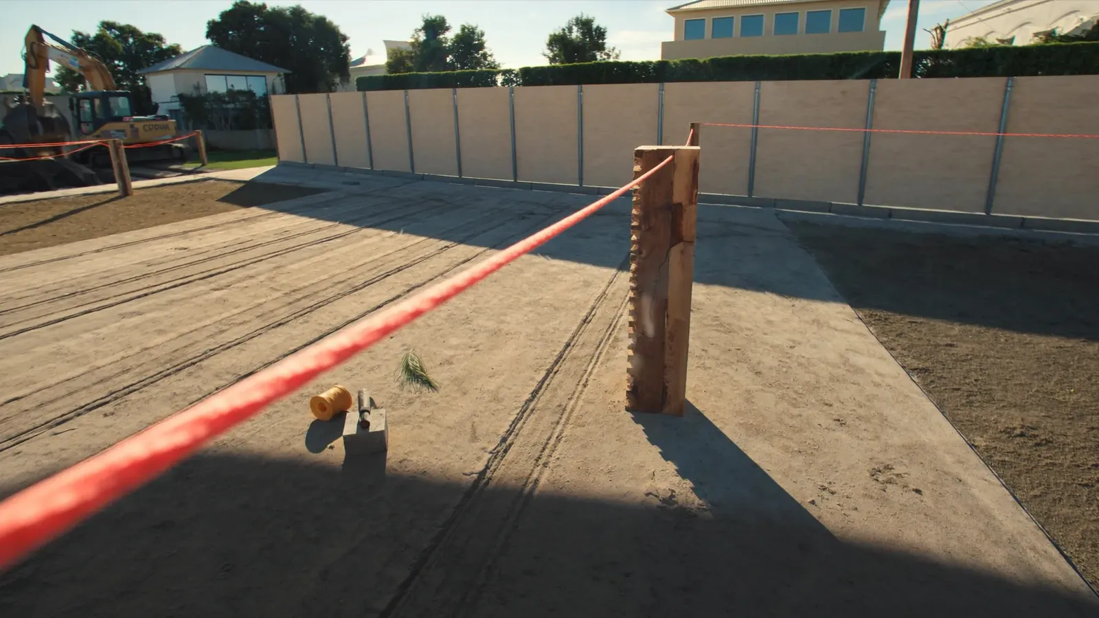 Setting out on a levelled Dubai villa plot: a taut string line running across the frame from a timber corner profile, a plumb bob hanging over the mark, hoarding along the boundary