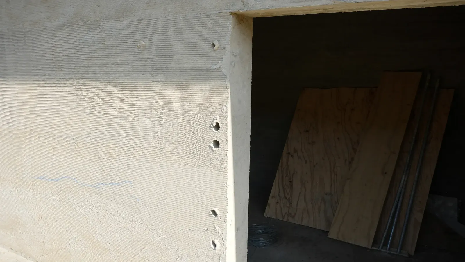 Stripped fair faced concrete on a villa frame, a vertical row of tie holes and horizontal board marks legible across the face, formwork panels stacked in the opening beyond