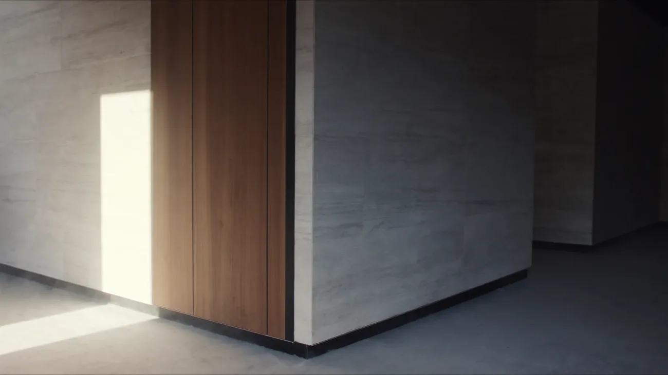 A finished villa interior: a full height run of walnut joinery meeting a stone clad wall on a corner, a shadow gap running true down each edge and a recessed shadow line at the floor, one block of hard sun on the stone