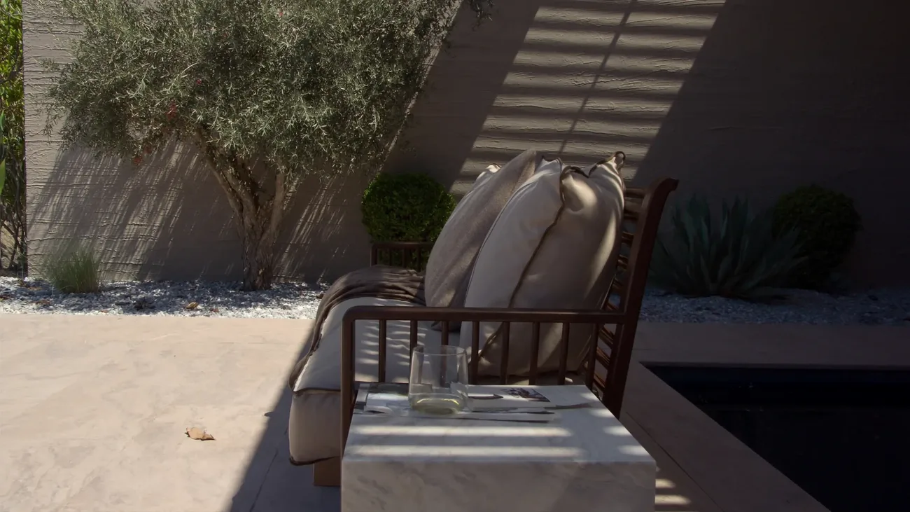 A shaded villa terrace: a bronze framed outdoor sofa with piped linen cushions and a throw, a travertine table with a glass set down on it, slatted pergola shadow across everything and a dark stone lined pool beyond
