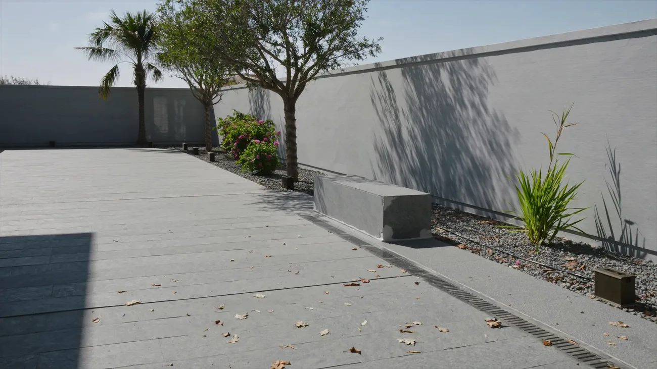 A villa courtyard in hard morning sun: large format limestone paving laid to a fall toward a stainless slot drain, a cast concrete bench, a gravel margin with drip irrigation, mixed planting against a rendered boundary wall and dry leaves blown across the stone