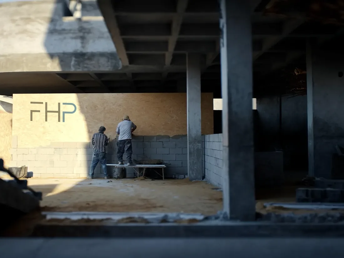 Two workers laying blockwork to a line on a villa wall, an FHP site hoarding standing behind them